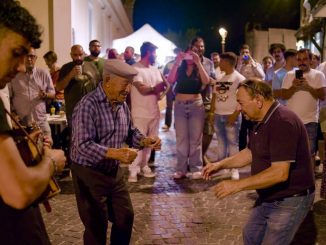 Felici & Conflenti, ballo notturno in un vicolo di pietra: un anziano con la coppola danza davanti a un altro uomo, accanto a un giovane che suona uno strumento a corde; intorno la gente in cerchio, qualcuno riprende col telefono.