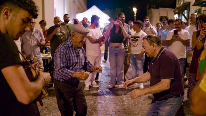 Felici & Conflenti, ballo notturno in un vicolo di pietra: un anziano con la coppola danza davanti a un altro uomo, accanto a un giovane che suona uno strumento a corde; intorno la gente in cerchio, qualcuno riprende col telefono.