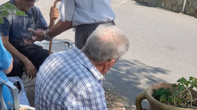Tre anziani conversano seduti all'ombra in un angolo di piazza a Martirano Lombardo, con alle spalle una casa dal tetto in tegole rosse